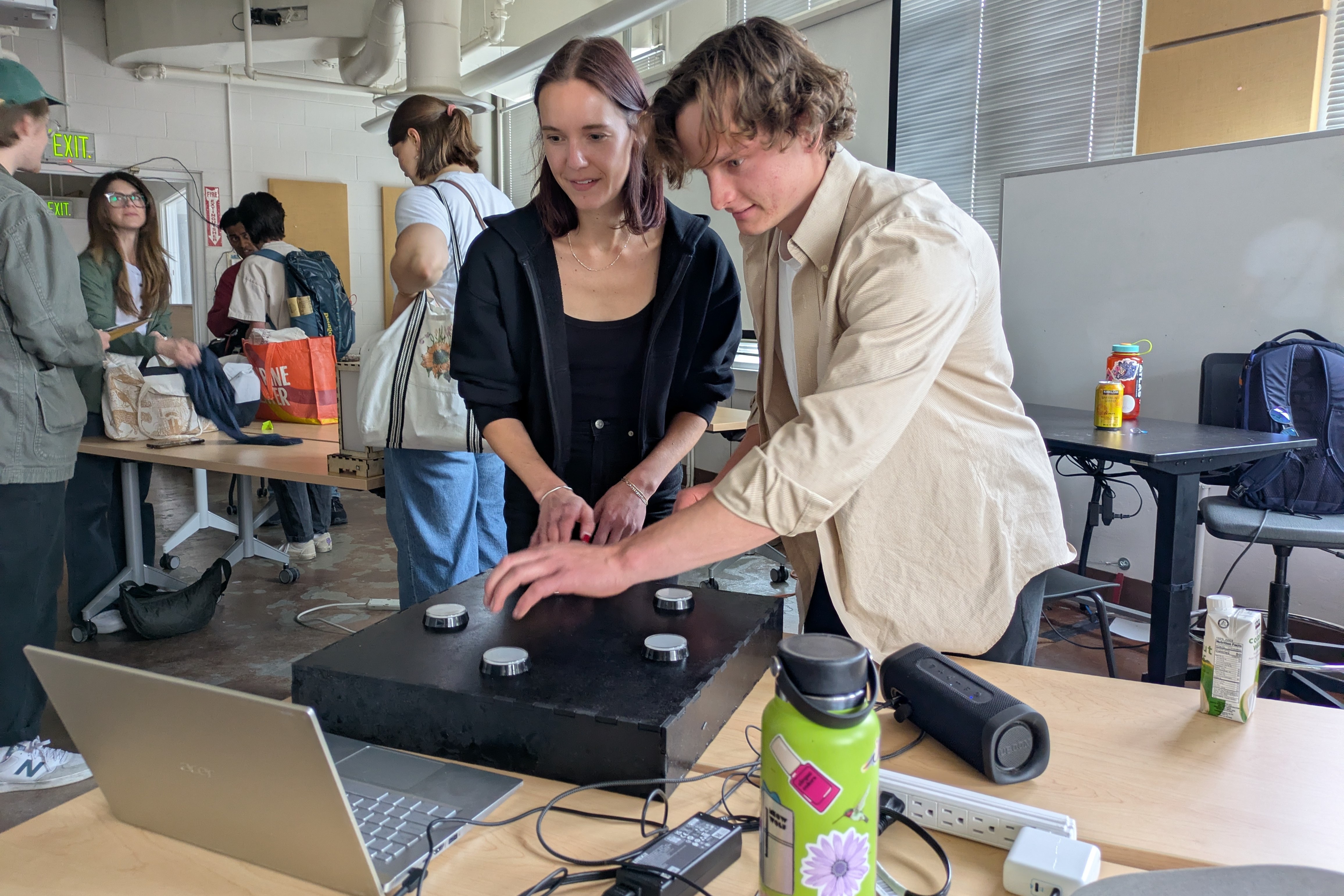 Two grad students, one male presenting and one female presenting, play a collaborative handmade video game together in a bright classroom. They are concentrating on the game, but smiling at the same time.