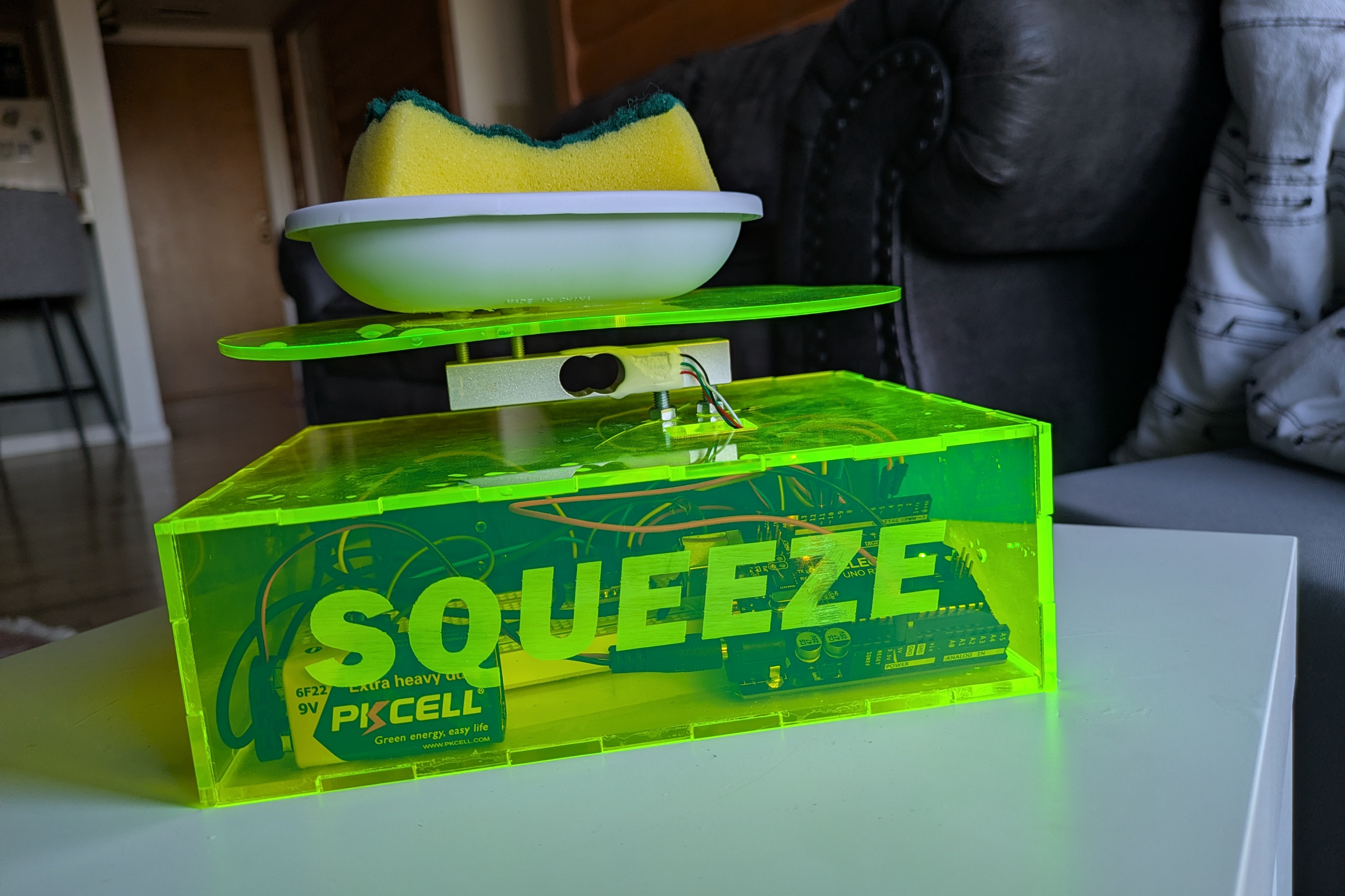 a clear box made of neon yellow laser cut 
        acrylic, with electronic parts and wires inside. on top of the box rests a soap dish on top of a weight sensor, with a sponge resting 
        on it. 